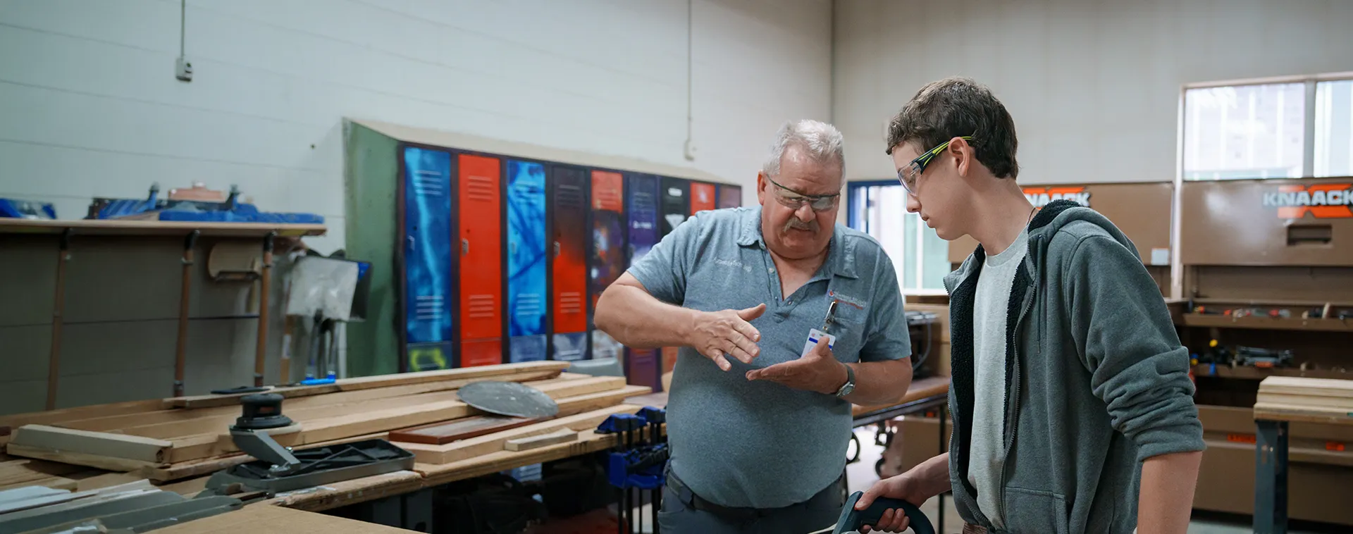 Instructor gesturing with hands while guiding student beside wood planks and lockers in workshop