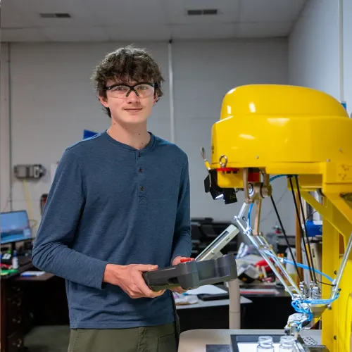 Student wearing safety glasses holds controller next to yellow robotic arm over worktable with components in lab