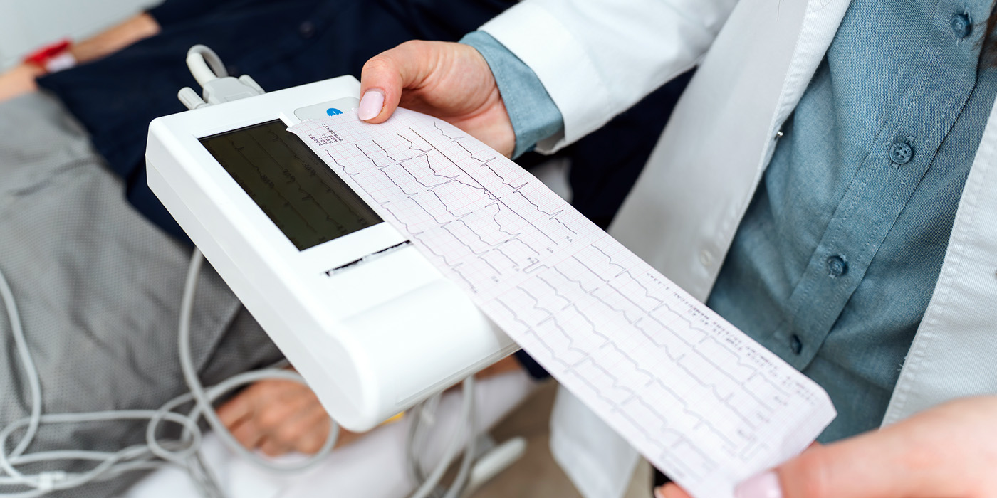Healthcare worker holding a portable EKG device printing a heart rhythm strip, with patient lying in background and wires attached