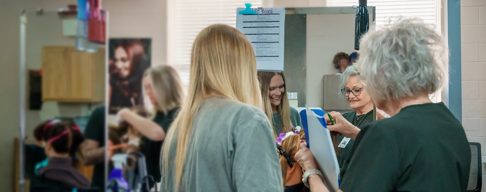 Instructor with clipboard reviewing work beside mirrors as student styles mannequin head with rollers in salon classroom
