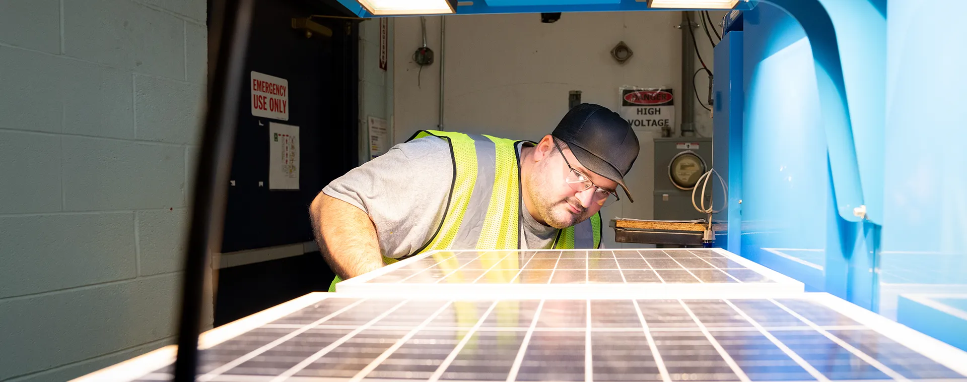 Person wearing safety vest inspects solar panel inside lab near equipment with &ldquo;Emergency Use Only&rdquo; and &ldquo;High Voltage&rdquo; signs visible