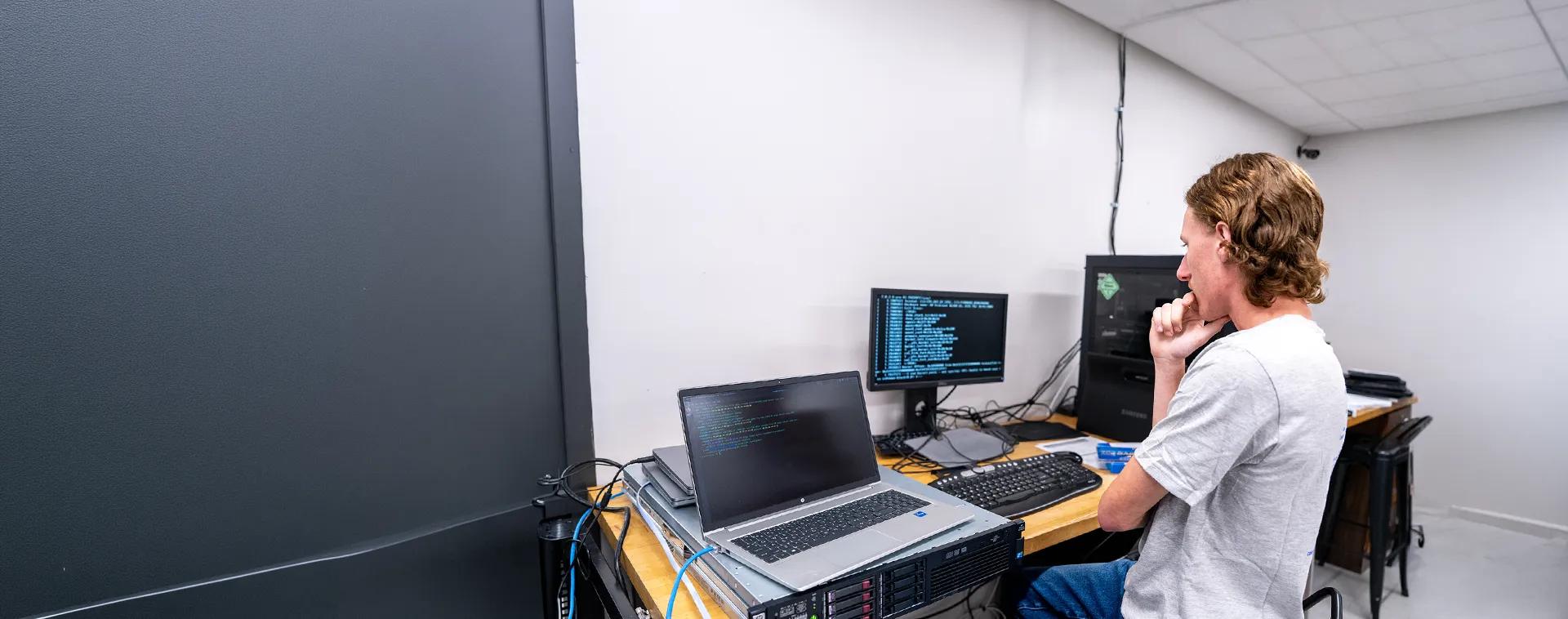Person seated at workstation using a laptop and monitors connected to server equipment in a computer lab.