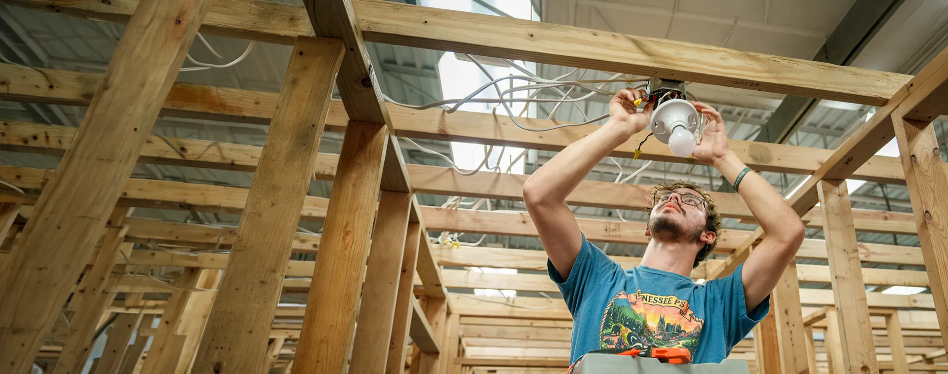 Student installing a light fixture and wiring within a wooden-framed electrical training structure.