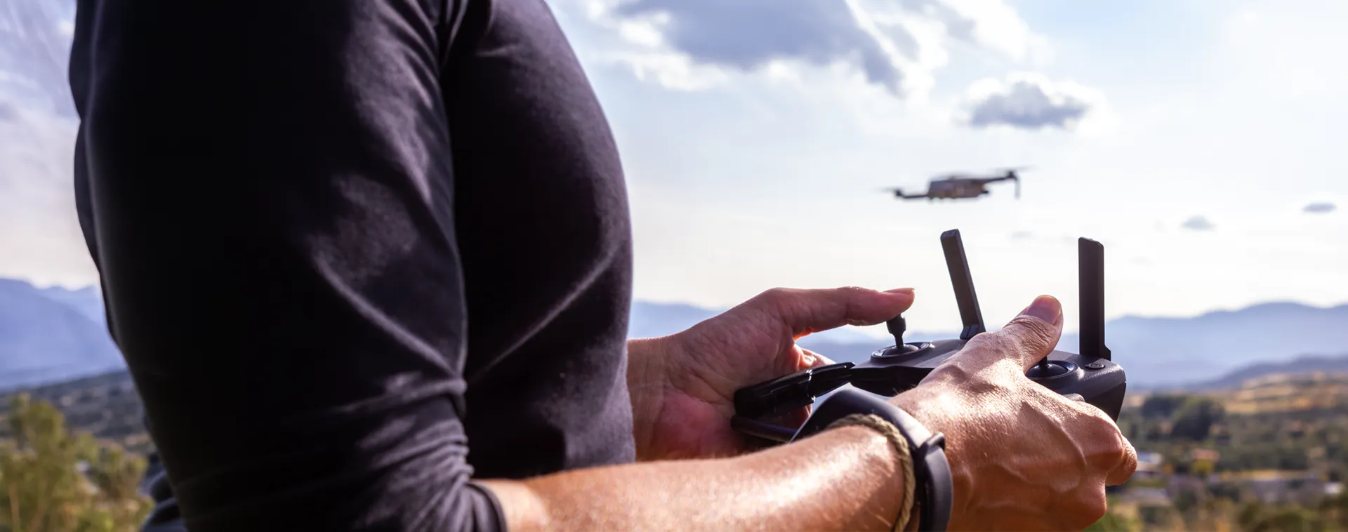 Hands operating a drone controller outdoors with a drone flying over a mountain landscape.
