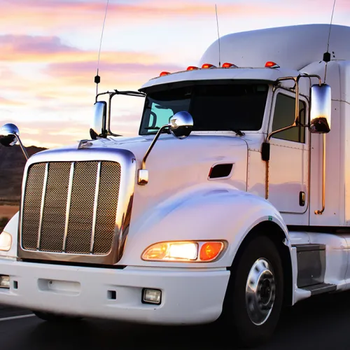 White semi-truck pulling a long trailer driving on a highway with mountains and a colorful sunset sky in the background