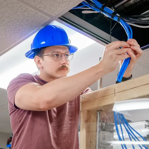 Person wearing a blue hard hat organizing blue network cables in an open ceiling cable tray.