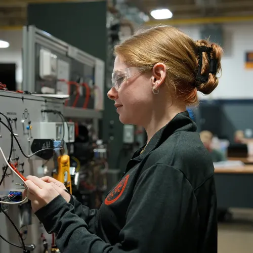 Student wearing safety glasses connecting wires on an HVAC control training panel.