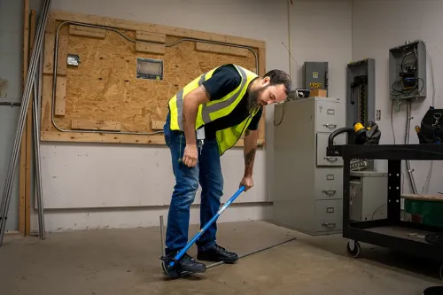 Person in safety vest bends conduit pipe using a manual bender in workshop with tools, wiring board, and cabinets in background