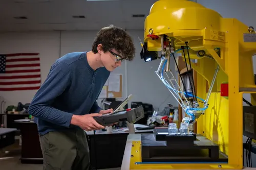 Student holding controller, operating yellow robotic arm placing items on platform in lab with American flag in background