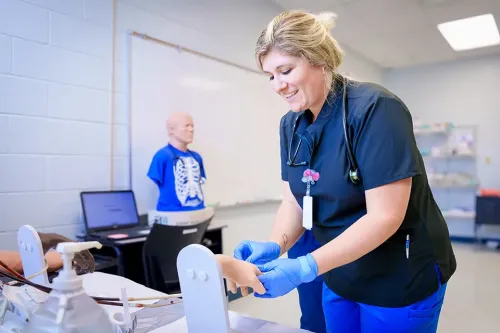 Student wearing gloves practices a medical skill on a training arm at a lab station with mannequin and computer in background