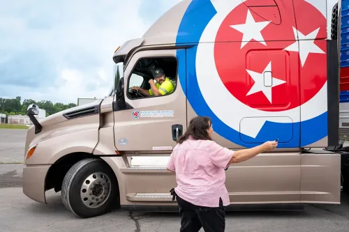 Person standing beside truck cab handing item to driver wearing safety vest next to Tennessee logo on door