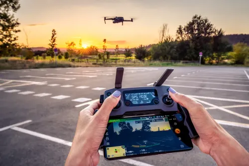 Drone controller with attached screen displaying flight data as a drone hovers above a parking lot at sunset.